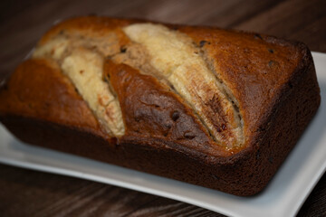 A freshly baked banana bread gateau is displayed on a white plate, showcasing its golden-brown crust and ripe banana slices on top. Perfect for a sweet treat or dessert.