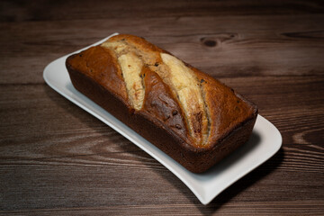 A freshly baked banana bread gateau is displayed on a white plate, showcasing its golden-brown crust and ripe banana slices on top. Perfect for a sweet treat or dessert.