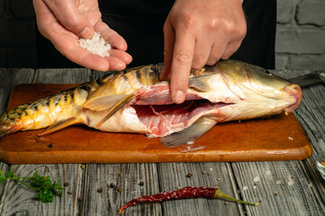 Hands expertly season a fresh fish on a wooden cutting board. Salt and a red chili pepper add flavor as the rustic kitchen enhances the culinary scene