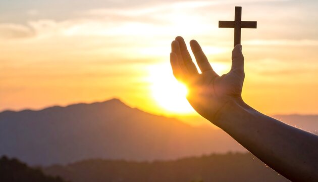 A hand holds a wooden cross against the sunset sky, silhouetted with mountain in the background. A visual depiction of faith and spirituality