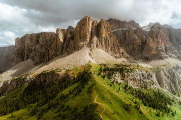 A scenic hiking trail leads along a lush green ridge toward towering rocky cliffs in the Dolomites. The dramatic light and cloudy sky create a moody atmosphere.