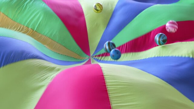 Colorful balls bounce on a vibrant parachute fabric during a fun group activity, creating dynamic motion and playful energy. Ideal for kids&rsquo; games and events.
