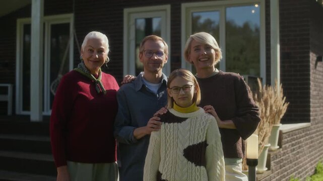 Zoom in portrait of happy Caucasian family members including senior grandmother, middle-aged parents and teenage daughter, posing for camera outdoors in front of modern house on sunny autumn day