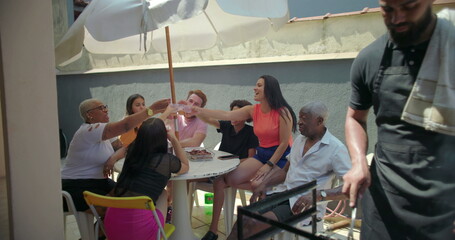 Group of diverse friends and family raising glasses in toast at outdoor barbecue, celebrating unity and joy around a table under a large umbrella in a vibrant backyard