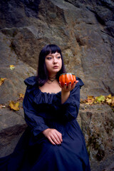 Obraz premium woman in a black long dress sits on black rocks on Halloween holding a pumpkin-shaped cup, a modern witch
