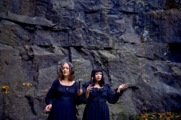 two women in the black long dresses near black rocks on Halloween holding a burning candle, performing a witch ritual, modern witch