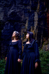 Naklejka premium two women in the black long dresses sitting on black rocks on Halloween, performing a witch ritual, modern witch