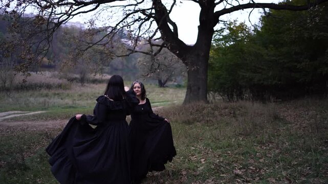      Two women in long black dresses perform a historical reenactment of the Edwardian era dancing lovers on a hill near a majestic oak tree for Halloween.