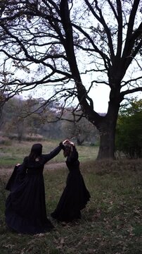      Two women in long black dresses perform a historical reenactment of the Edwardian era dancing lovers on a hill near a majestic oak tree for Halloween.