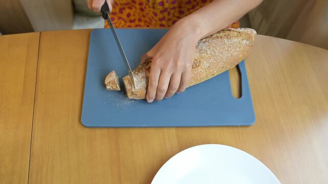 Close-up video of a woman slicing fresh multigrain bread with a knife on a cutting board at the kitchen table, preparing food at home