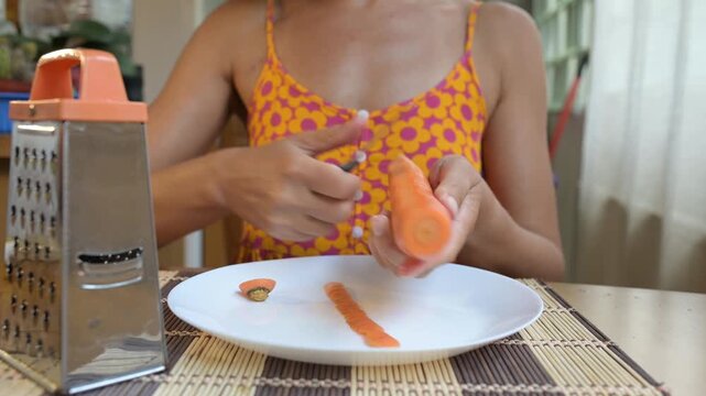 Close-up video of a woman peeling a fresh carrot with a knife over a plate at the kitchen table, preparing vegetables for cooking