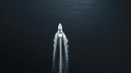 Aerial top-down photo of a white motorboat cruising through deep dark blue water, leaving a dynamic V-shaped wake behind. The minimalistic composition emphasizes the contrast between the boat and the