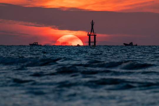 View of a fiery sunset bleeding across the sky, silhouetting boats and a nautical marker, waves ripple in the foreground, Berni&Atilde;&uml;res-sur-Mer, Normandy, France.