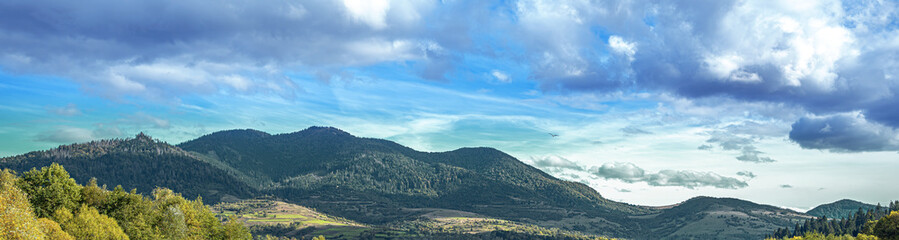 Fototapeta premium A panoramic view of green forested mountains and a valley under a dramatic cloudy sky in a peaceful natural landscape.