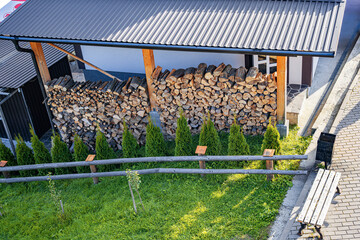 Neatly stacked firewood under a shed roof, with a low wooden fence separating the woodpile and...