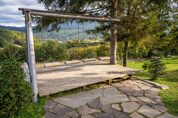 A rustic wooden swing set on a platform overlooks a scenic valley with mountains, forests, and a village in the distance on a sunny day.