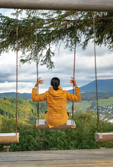 A woman in a yellow jacket sits on a swing overlooking a scenic mountain valley with forests, villages, and cloudy skies, creating a peaceful outdoor travel scene.