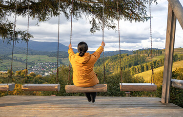 A woman in a yellow jacket sits on a swing overlooking a scenic mountain valley with forests, villages, and cloudy skies, creating a peaceful outdoor travel scene.
