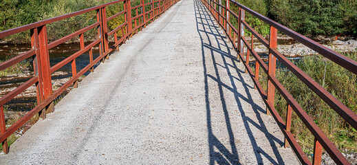 A straight, narrow concrete bridge with rusty red metal railings, as it spans a river surrounded by overgrown greenery.