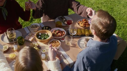 High angle slowmo shot of united Caucasian family holding hands joining together in prayer while sitting around dining table during family celebration on lovely sunny day outdoors - Powered by Adobe