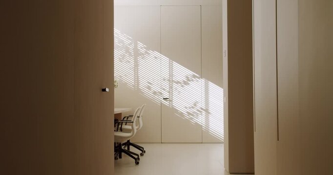 Sunlight streams through window blinds, casting shadows in a modern, minimalist office space. White chairs sit beside a simple table, Natural light illuminates the room, work