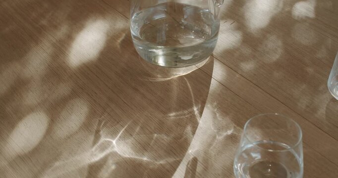 Sunlight streams through a window, casting dancing shadows on a wooden table where a glass pitcher of water sits beside two clear glasses. One glass is empty, the other half full,