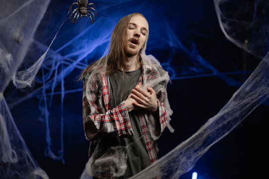 Creepy scary zombie man with ugly makeup holding his chest has panic attack feels severe pain standing on spooky spider web background. Halloween concept. Horror film.