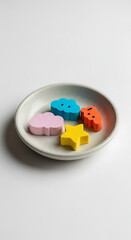 Overhead shot of a white plate with colorful wooden shapes, including a pink cloud, blue cloud, yellow star, and orange strawberry, isolated on white background these playful shapes create a cheerful 