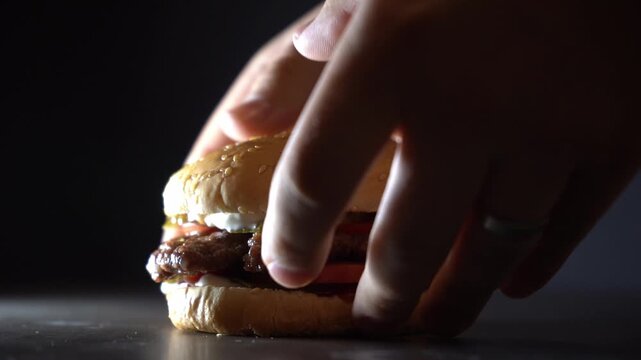 Hand picking up a gourmet burger from a table under dramatic light, capturing motion, food texture, and cinematic culinary atmosphere in a dark minimal scene.