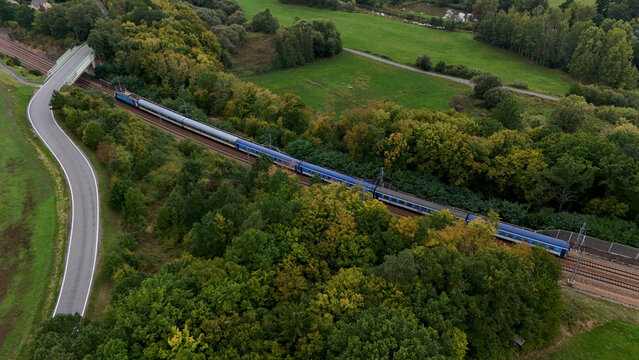 Aerial view of passenger train moving through green countryside with winding road and fields, modern rail transport integrated into rural landscape and sustainable mobility concept.