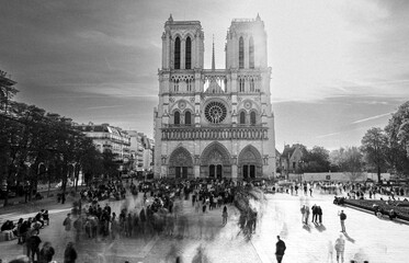 A lively scene unfolds as people gather near Notre-Dame-de-Paris at sunset. The historic cathedral's architecture is beautifully highlighted in black and white, capturing the moment.