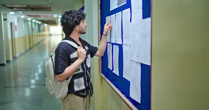 Indian young adult gen z upset sad pupil boy look notice do check exam results study report campus indoor area. Stress male man feel bad wear bag read new paper list data notice inside college place