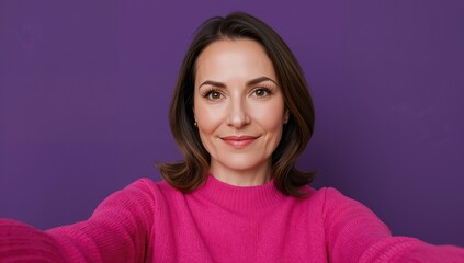 Fototapeta premium Mature woman taking a selfie wearing a bright pink jumper against a solid purple background with a warm smile and shoulder-length brunette hair