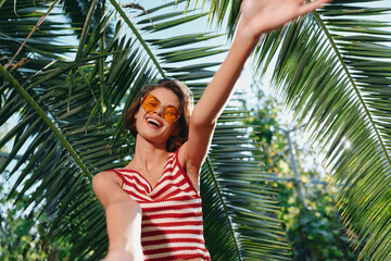 A cheerful woman wearing a striped top and sunglasses takes a sunny selfie under palm fronds, smiling broadly. The vibrant outdoors evokes carefree summer vibes and a lively fashion moment.