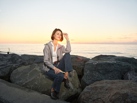 A cheerful woman sits on rocky shore at sunset, wearing a casual jacket and jeans. She smiles softly toward the horizon, creating a calm, peaceful coastal portrait.