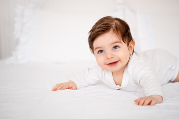 A baby under a blanket on a white bed at home, a place for text, a cute smiling little child getting ready for bed