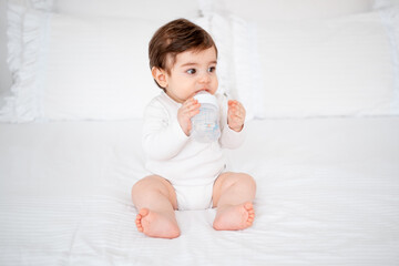 baby with a bottle of milk or water on a white bed at home, baby food feeding concept, place for text, cute smiling little baby
