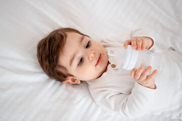 baby with a bottle of milk or water on a white bed at home, baby food feeding concept, place for text, cute smiling little baby