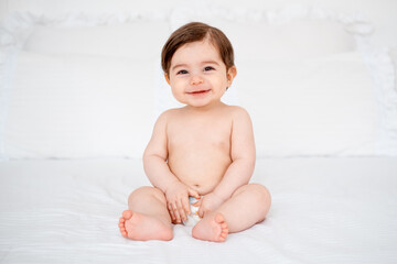 A baby in diapers on a white bed at home, a place for text, a cute smiling little child