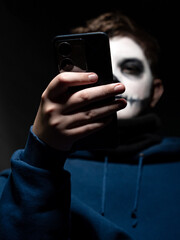 A young person with skull makeup, holding a smartphone, pointing at its screen with their finger.