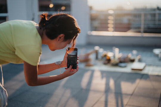 A young woman captures a beautiful food arrangement with her phone camera on a rooftop during sunset. The scene highlights creativity, outdoor leisure, and the pursuit of aesthetic enjoyment.