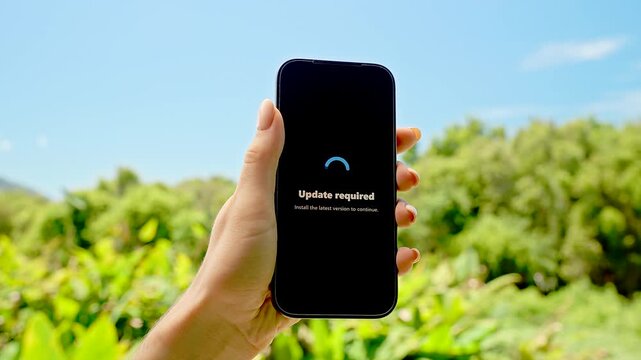 Smartphone in Hand Displaying Update Required Screen Against Sunny Sky and Green Grass Background, Concept Technology Maintenance and System Refresh