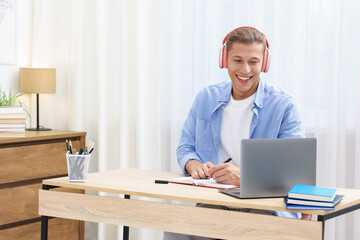 Fototapeta premium Man in headphones taking notes while passing online test via laptop at table indoors