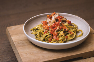 Tagliatelle pasta tossed in pesto, topped with sliced meat and chunky diced red tomatoes, served in a ceramic bowl on wood.