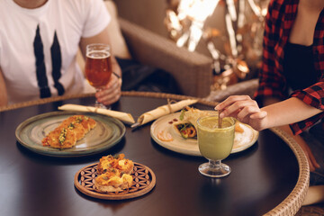 Close-up of friends enjoying a casual, healthy meal and drinks on a patio, featuring a green smoothie, craft beer, and gourmet appetizers on a dark table.