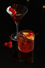 Two elegant, classic red cocktails (Manhattan, Old Fashioned, or Martini) with maraschino cherry and orange garnishes, set dramatically on a black reflective background.