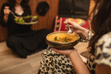 Close-up of woman holding rustic bowl with creamy pumpkin soup, garnished with herbs, while dining...