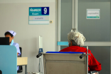 An elderly woman with gray hair in a red dress sits in a wheelchair and waits in a hospital examination room with a sign in Thai. A nurse stands behind her.