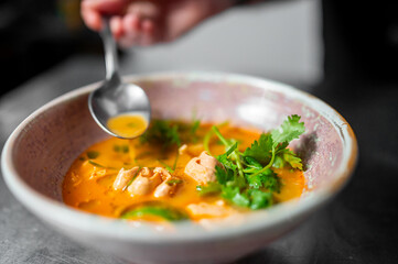 Close-up of Thai-style chicken soup with orange broth, cilantro, and green chili in a rustic bowl. A hand holds a spoon, capturing a moment of tasting. Warm, vibrant, and appetizing