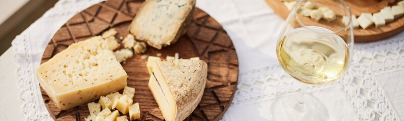 Website header shot of assorted cheese wedges and cubes arranged on wooden platter beside glass of wine on lace tablecloth, showcasing gourmet food selection for tasting or entertaining